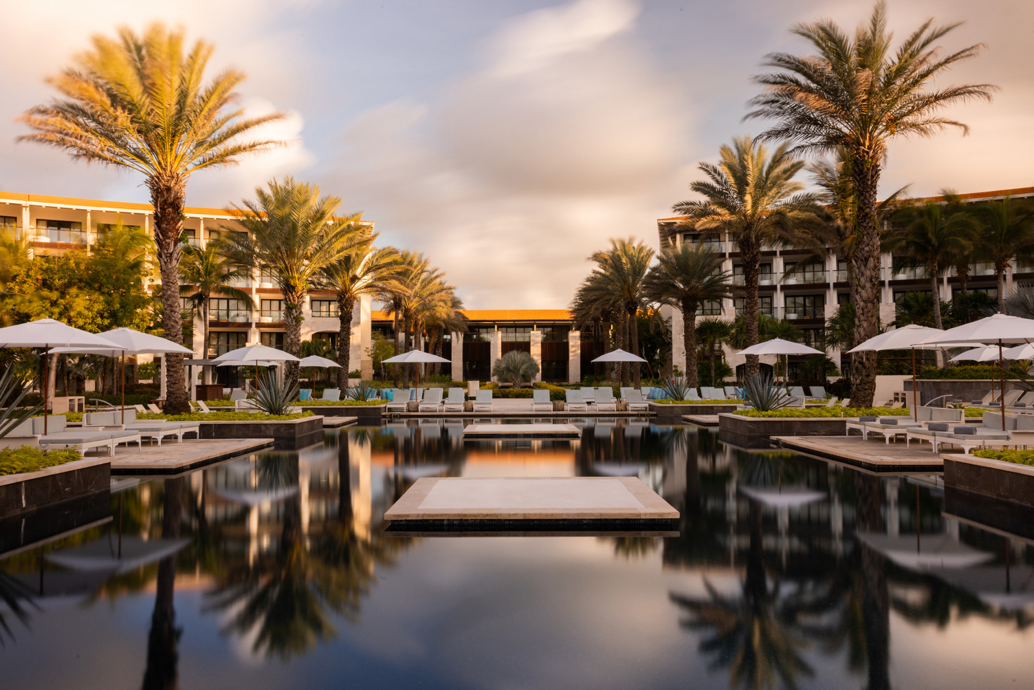 Área de alberca del resort con palmeras, camastros y sombrillas al atardecer, reflejándose en el agu