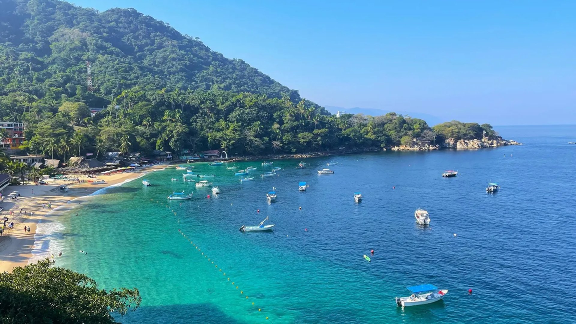 Coastal view with clear turquoise water, small boats, lush green mountains, and a sandy beach on a sunny day.