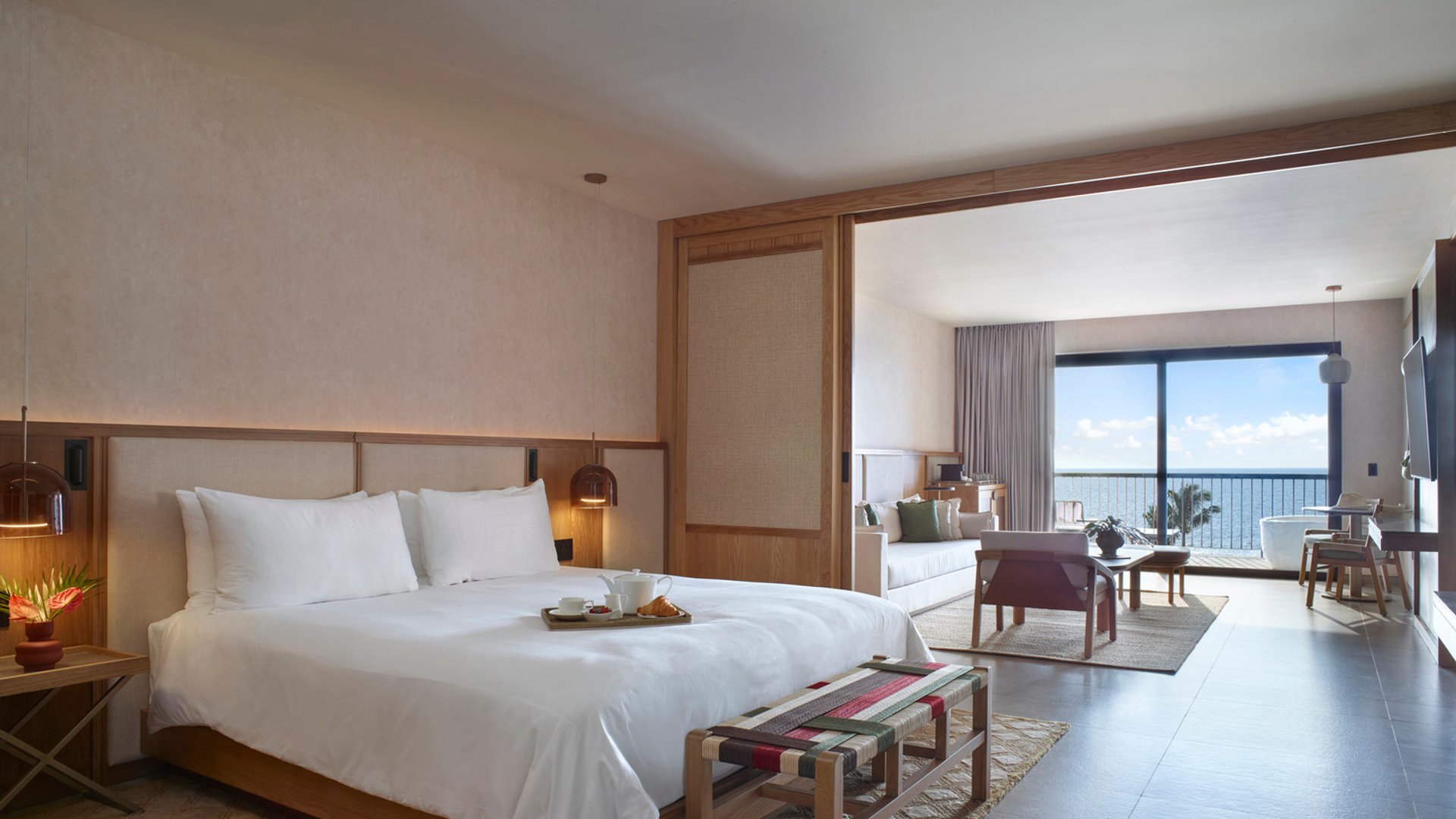 Luxurious suite with a king bed, ocean view, modern decor, sitting area, and sliding glass doors opening to a balcony.